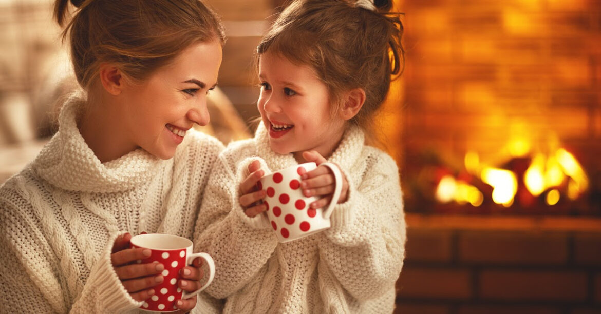 A woman and her daughter enjoy a warm beverage by the fireplace